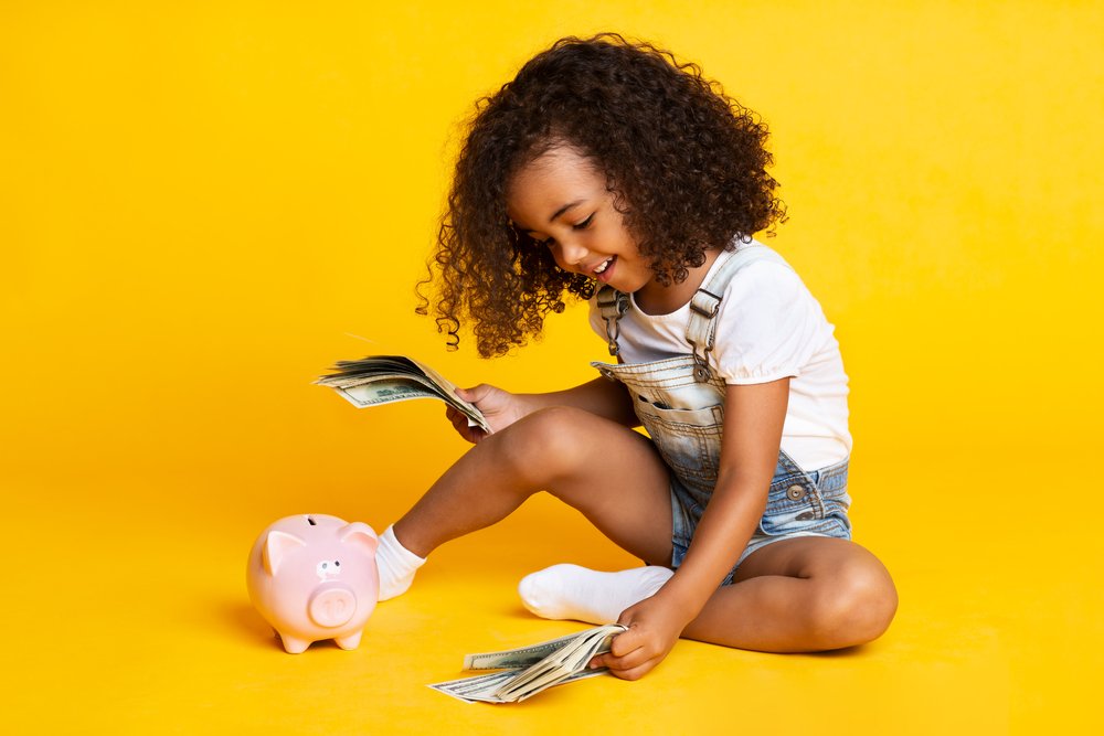 Little girl playing with money, piggy bank nearby on yellow background - Traveling with grandkids - Adventures in NanaLand
